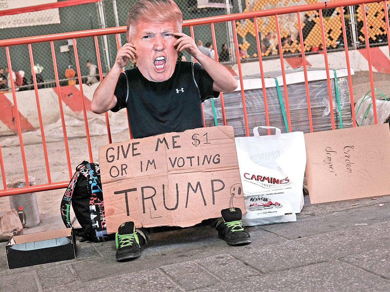 شخص يرتدي قناعاً لوجه ترامب وأمامه لافتة كتب عليها: «ادفع لي دولاراً وإلا فسأصوِّت لمصلحة ترامب»، في نيويورك أمس الأول (أ ف ب)