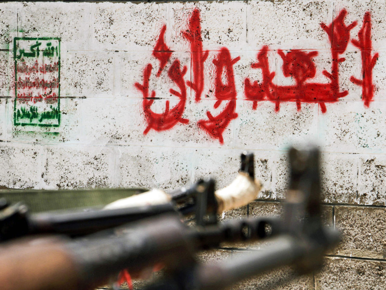 مقاتل يمني موال للرئيس اليمني يصوّب بندقية كلاشنيكوف نحو جدار يحمل شعار الحوثيين.