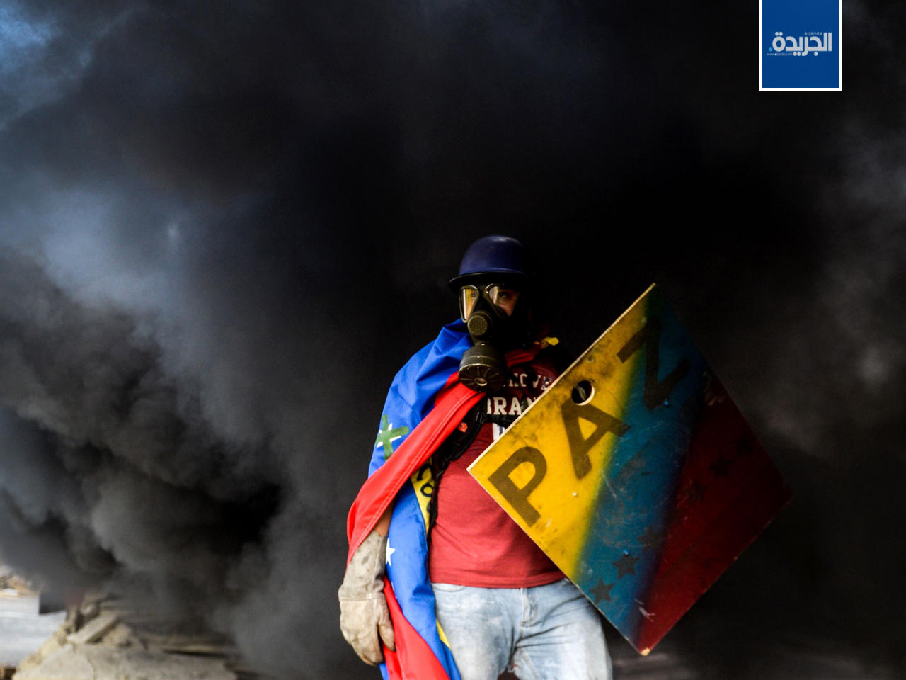 معارض فنزويلي يرتدي قناعاً غازياً ويحمل درعاً بينما يلف حوله دخان شاحنة اشتعلت فيها النيران