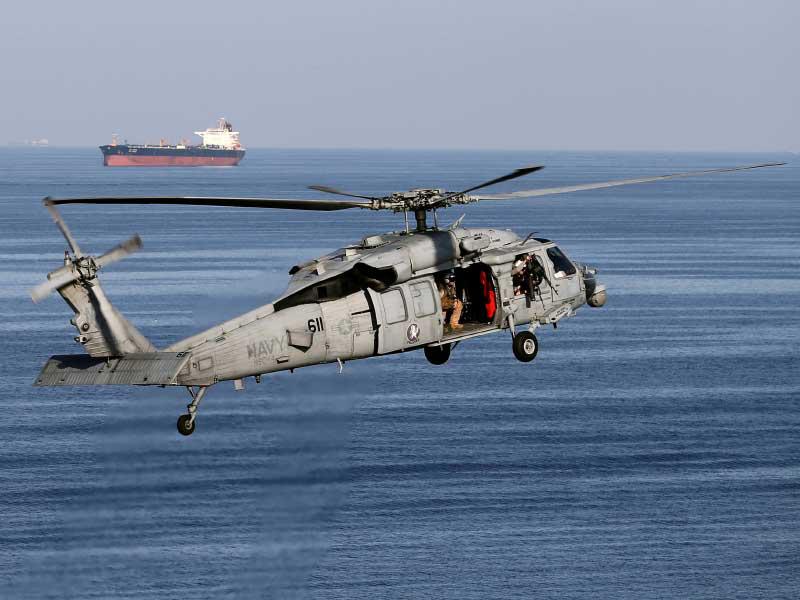 طائرة هليكوبتر من طراز تحلق في الهواء مع ناقلة نفط في الخلفية بينما تشق حاملة الطائرات الأمريكية جون سي ستينيس الطريق إلى الخليج عبر مضيق هرمز