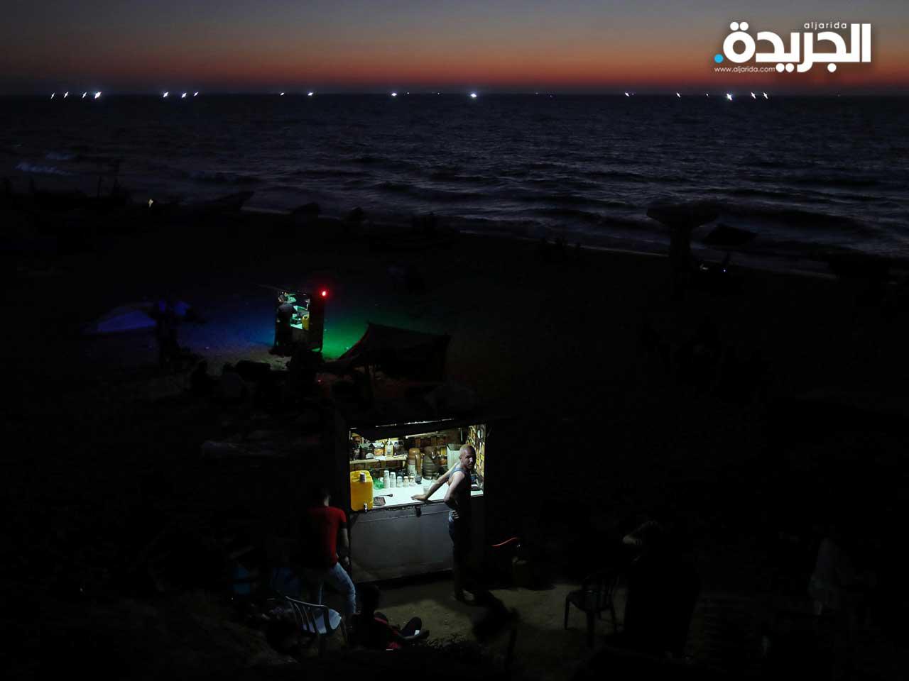 بائع فلسطيني يبيع القهوة والشاي أثناء انقطاع التيار الكهربائي وهو يقف بجانب كشكه مستخدماً أضواء تعمل بالبطارية ، على شاطئ في شمال قطاع غزة