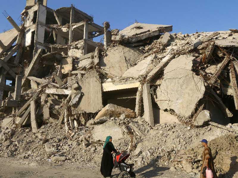 امرأة تدفع عربة أطفال وهي تسير خلف أنقاض المباني المتضررة في الرقة