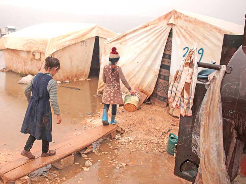 طفلتان نازحتان من مدينة إدلب السورية تعبران إلى خمية أسرتهما وسط الوحل على حدود تركيا  (أ ف ب)