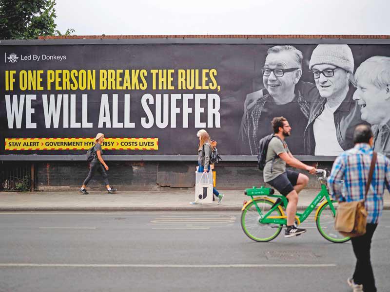 بريطانيون يمرّون أمام لافتة كبيرة في لندن عليها صورة جونسون والمستشارين دومينيك كيمينغز وميكايل غوف كتب عليها «اذا خرق أحدهم القانون فسنعاني جميعنا» (ا ف ب)