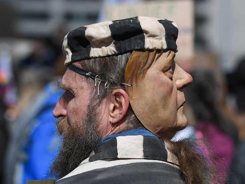 ألماني يضع قناعاً لميركل خلال تظاهرة ضد إجراءات «كورونا» في مدينة شتوتغارت أمس (رويترز)