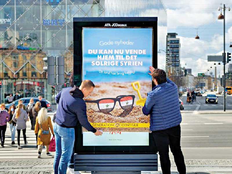 دنماركيان مناهضان للمهاجرين يلصقان إعلاناً في كوبنهاغن كتب عليه: «بشرى سارة يمكنك العودة إلى سورية المشمسة. بلدك بحاجة إليك».n