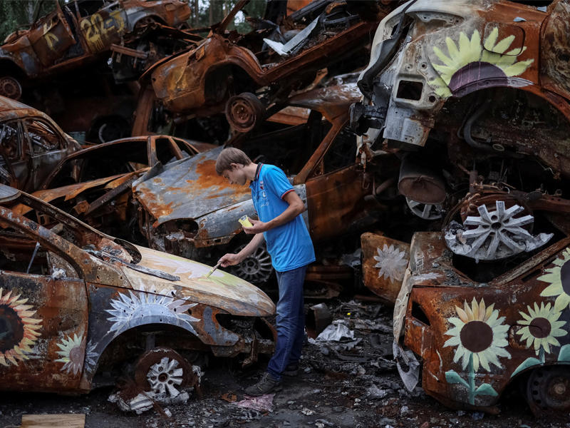 شاب أوكراني يرسم على سيارات محترقة بفعل القصف الروسي في بلدة أربين بكييف (رويترز)